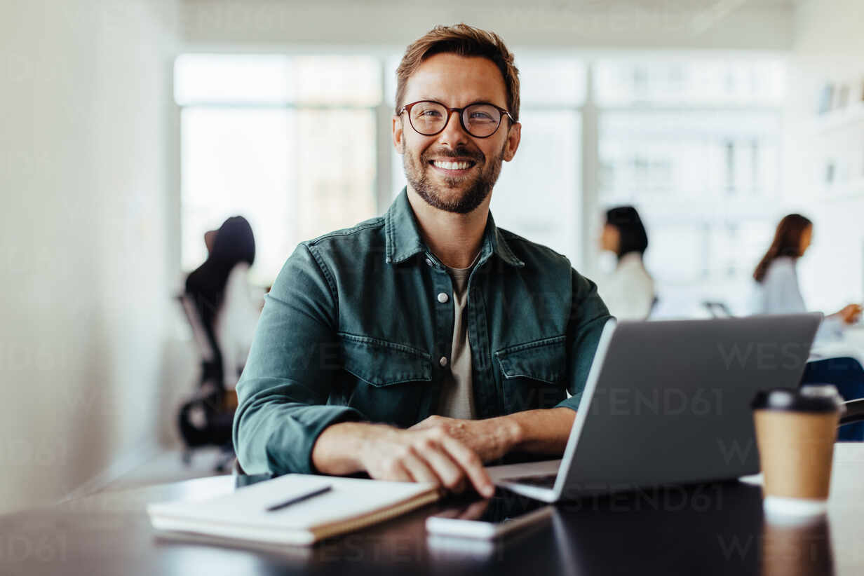 portrait-of-a-business-man-sitting-in-an-office-with-his-colleagues-in-the-background-happy-business-man-working-in-a-co-working-office-JLPSF28798